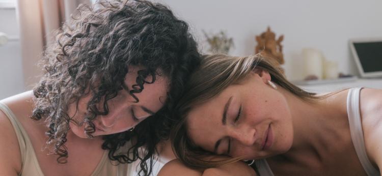 Two young women leaning on each other with eyes closed during a group yoga class with women of different age groups and body types. Shot in a beautiful bright and spacious shala with natural light. Muted color palette.