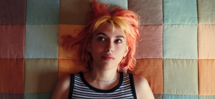 Thoughtful teenager with multicolored hair lying in bed over a multicolored quilt and looking away.