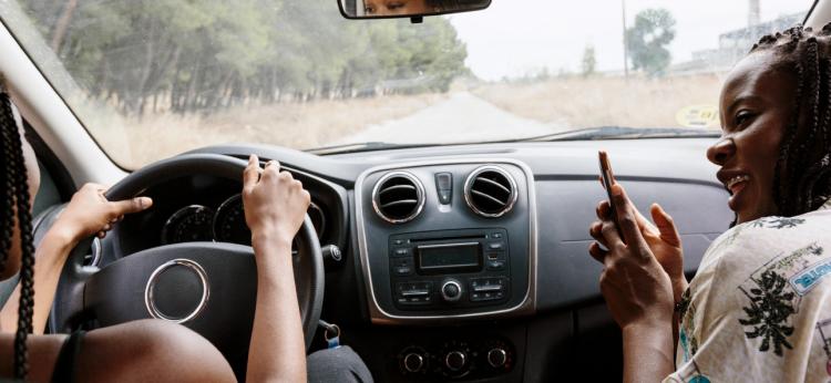 Two young attractive women inside a car. One of them taking a picture of the other on her smart phone. 