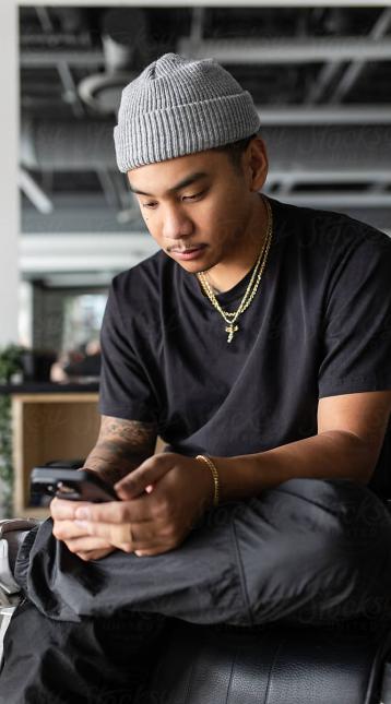 A young confident, filipino barber sits in his barber chair as he uses his cell phone.